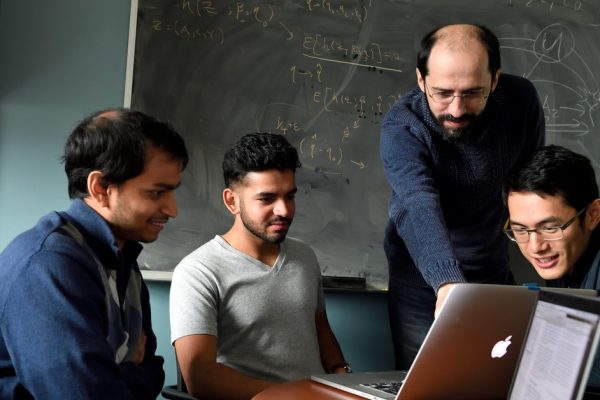 Ilya Schpitser and three students gather around a Macbook in front of a blackboard with mathematical equations written on it.