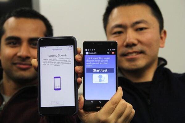Two researchers hold up smartphones displaying mobile applications for detecting Parkinson's disease. The phone on the left reads "Tapping Activity. Tapping Speed. Speed of finger tapping can reflect severity of motor symptoms in Parkinson disease. This activity measures your tapping speed for each hand. Your medical provider may measure this differently. Next." with a purple icon of a smartphone. The phone on the right reads "HopkinsPD. 1. Voice test. Find a quiet location. When you are ready, press the button below. Start test." with a purple icon of a stick figure holding up a phone.