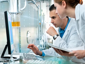 Stock image of two researchers in a lab wearing white coats and looking at a computer screen.