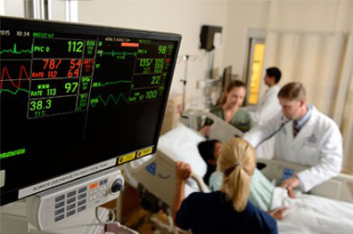 Doctors gather around a patient's bed in the background. In the foreground, a monitor displaying the patient's vital signs.