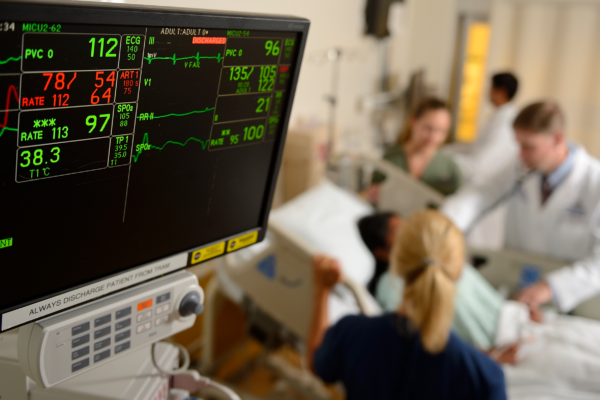 Doctors gather around a patient's bed in the background. In the foreground, a monitor displaying the patient's vital signs.