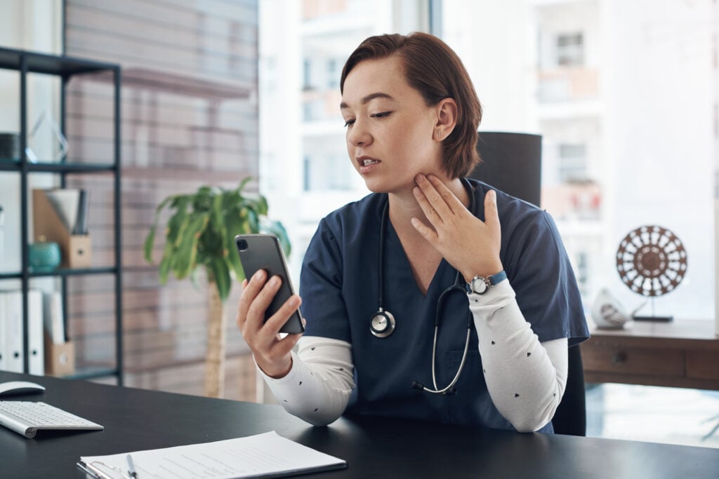 A young doctor in an office holds a cell phone and demonstrates feeling her throat.