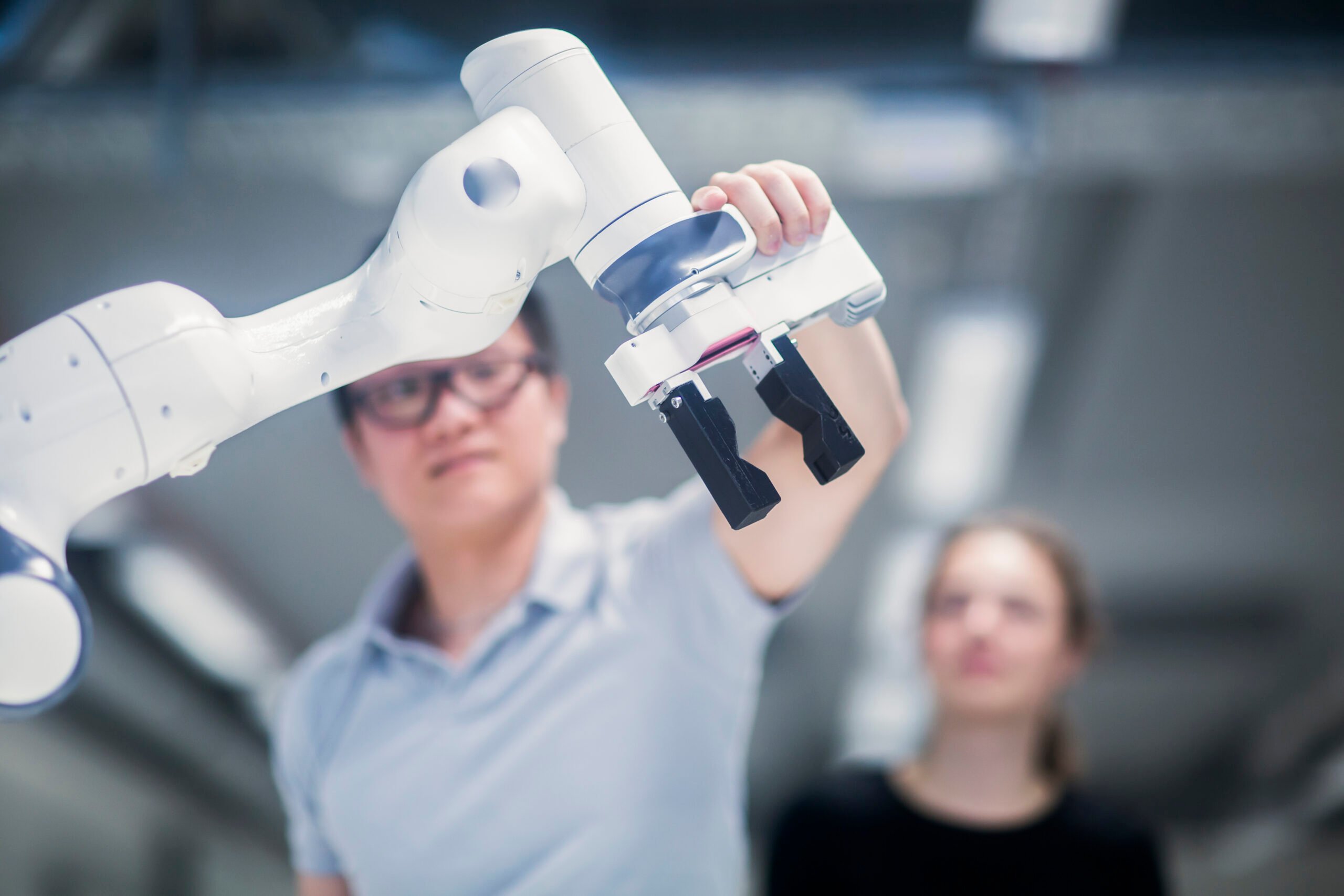 A student grasps a robotic arm's end effector as another student looks on.