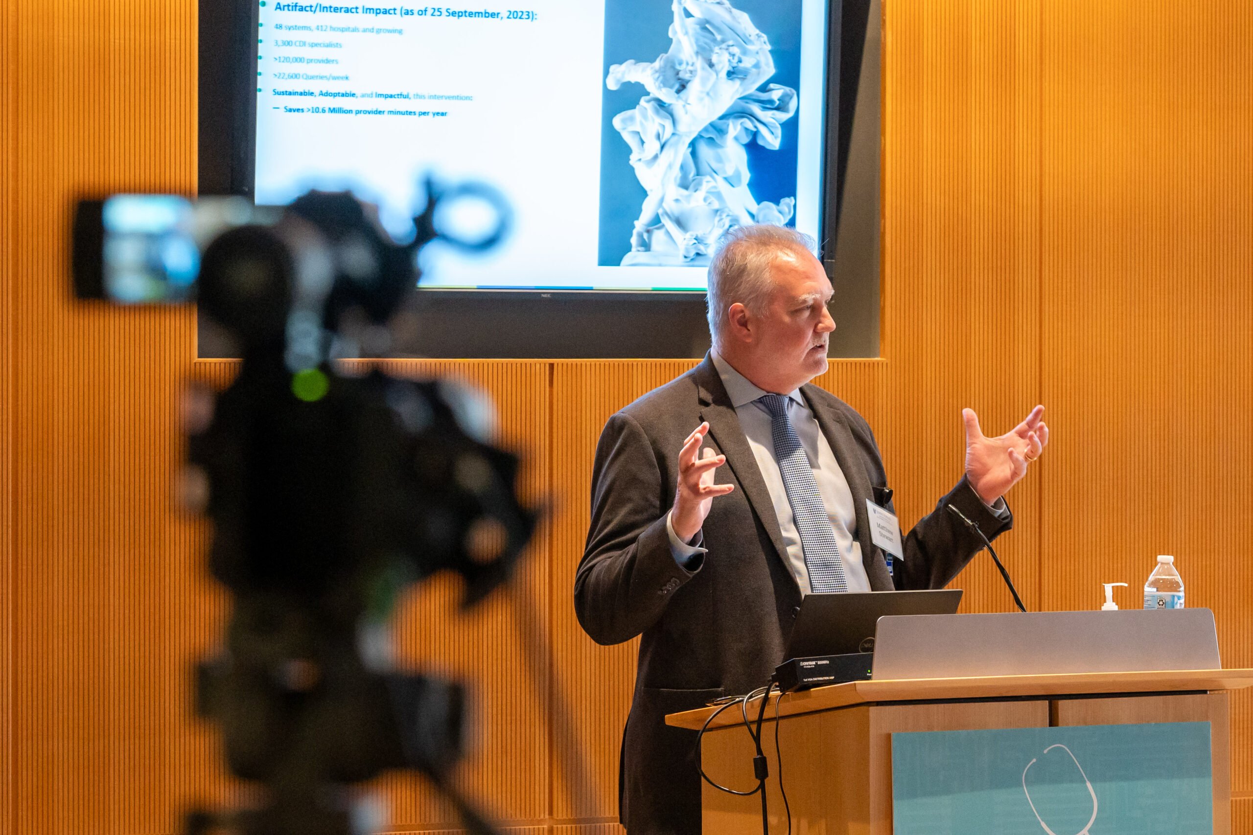 Matthew Stewart gestures at a podium with a camera in the foreground.