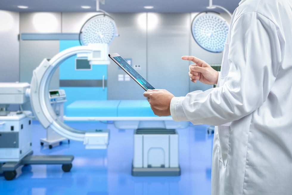A person in a white doctor's coat gestures to a tablet in their hand. Behind them is an operating room with a C-arm X-ray imaging machine.