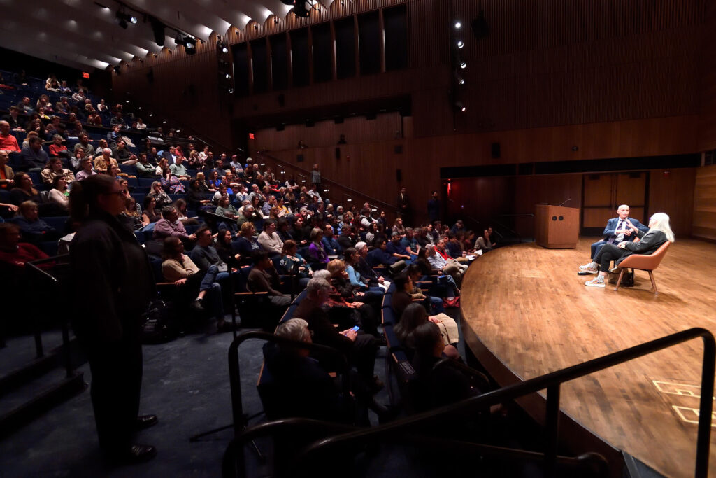 Two people sit on a stage and converse in front of a full auditorium audience.
