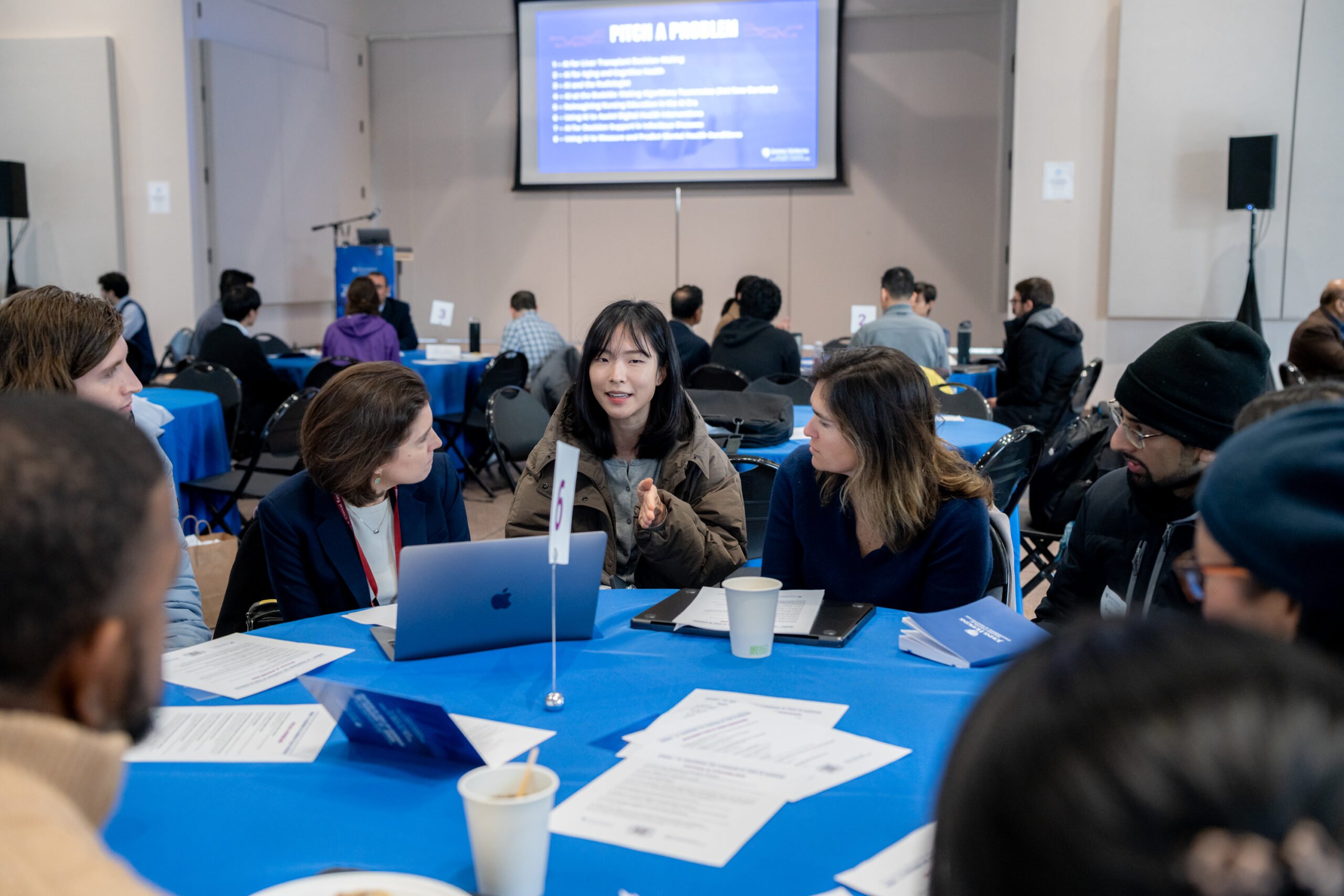 Malone_Symposium_25-26 Attendees sit around a round table with a number 6 card on it and discuss. "Pitch a Problem" and various topics are projected onto a screen behind them.