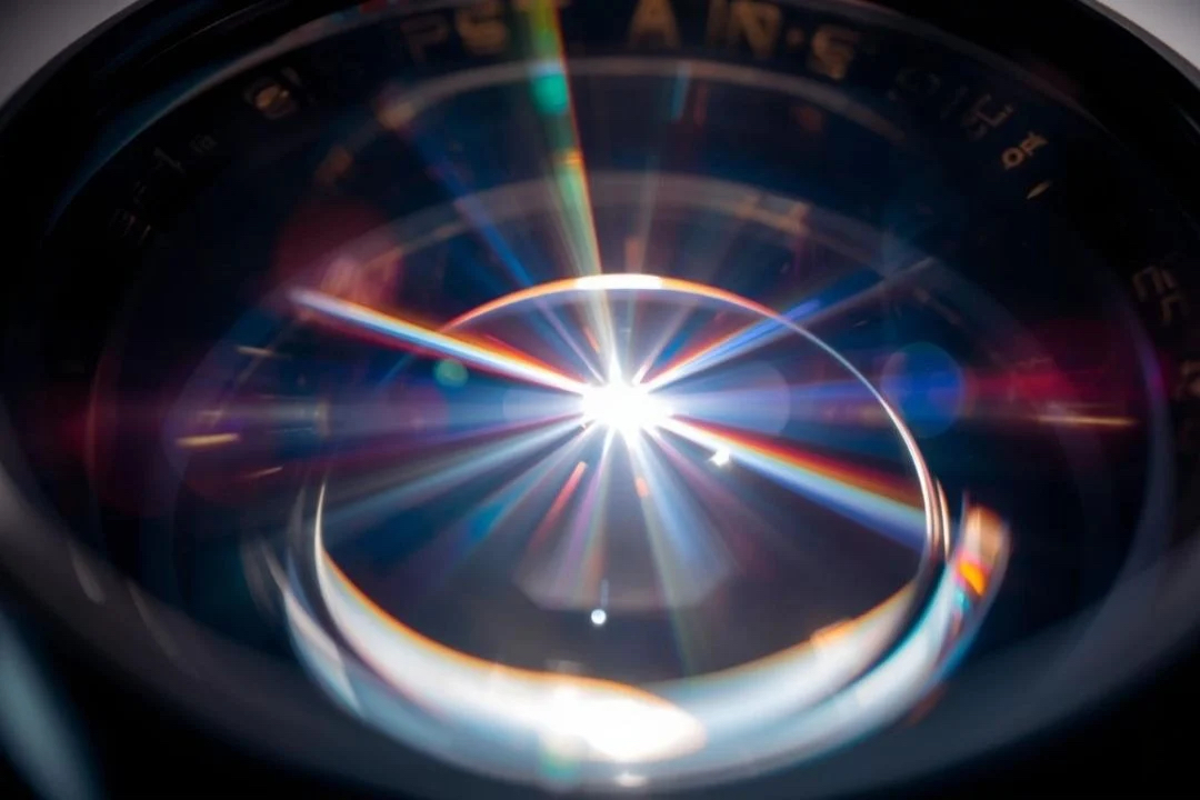 Extreme close-up of a camera lens capturing a brilliant, prismatic light flare with rainbow-colored rays radiating from the center.
