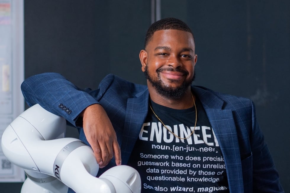 A. Michael West, Jr. leans against a robot arm wearing a suit jacket and a T-shirt displaying the definition of "engineer".